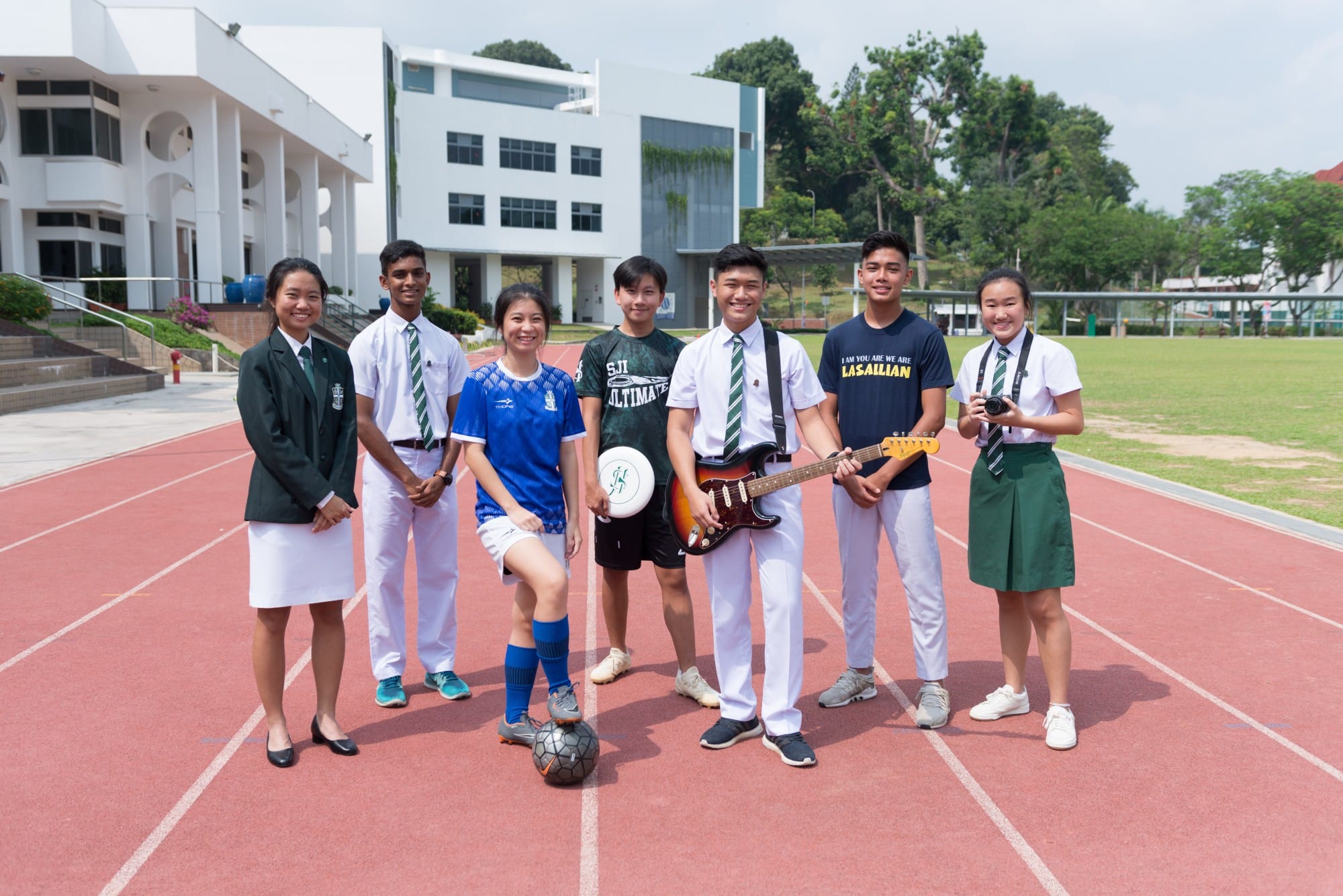 Seven students stand on a red running track, holding a guitar, frisbee, soccer ball, and camera, with school buildings behind.
